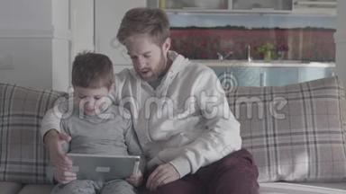 父子俩用数码平板电脑坐在大客厅的沙发上。 爸爸教他的孩子。 快乐的家庭开支